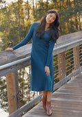 A woman in the Woolx Caroline Ribbed Twirl Dress in Majolica Blue and brown ankle boots stands on a wooden bridge, leaning on the railing with trees and sunlight in the background.