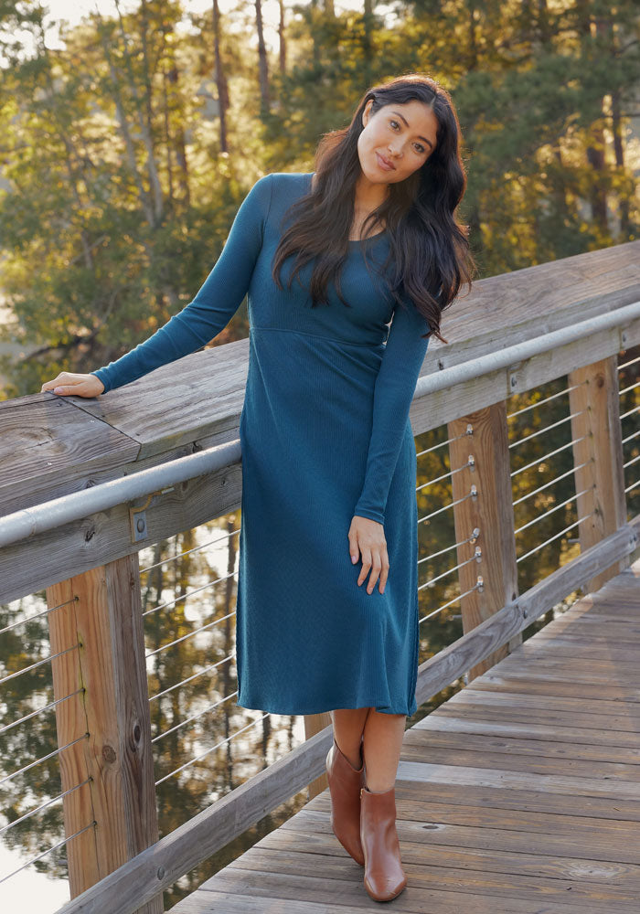A woman in the Woolx Caroline Ribbed Twirl Dress in Majolica Blue and brown ankle boots stands on a wooden bridge, leaning on the railing with trees and sunlight in the background.
