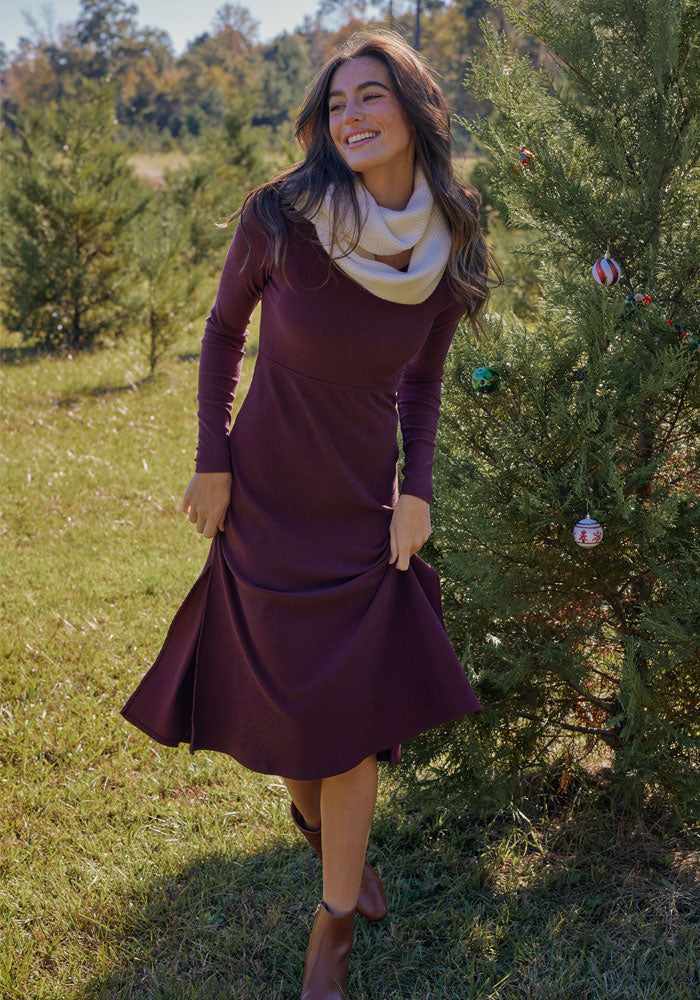 A woman smiles outdoors beside a decorated evergreen, wearing the Woolx Caroline Ribbed Twirl Dress in Toasted Garnet with a cream scarf and brown ankle boots on a grassy field with more trees behind her.