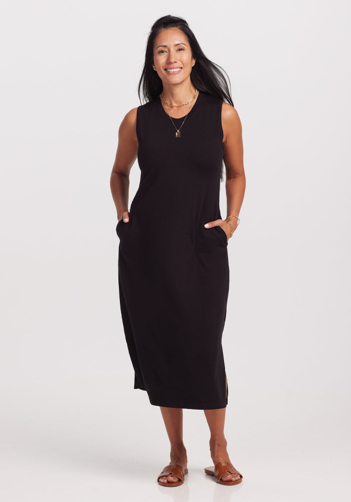 A woman with long dark hair smiles in a Woolx Cassie Dress - Black, featuring pockets. Her outfit is completed with bracelets, a necklace, and brown sandals. The plain white background enhances this perfect travel ensemble.
