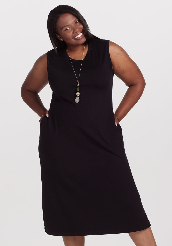 Against a plain white backdrop, a smiling woman confidently poses with hands on her hips, wearing the Woolx Cassie Dress in black. She accessorizes with a long necklace featuring three circular pendants. | Le’Quita is 5’11”, wearing a size XL