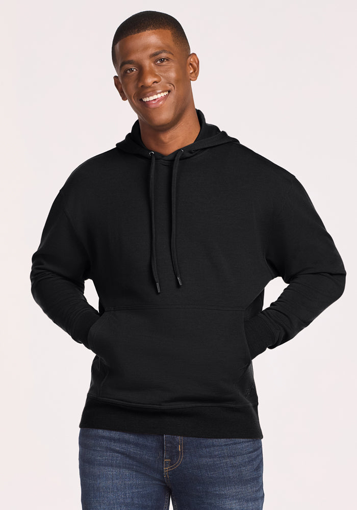 A young man smiles wearing the Woolx Chase Hoodie - Black, featuring moisture-wicking fabric and a front pocket, paired with blue jeans against a simple backdrop. | Trell is 6’2”, wearing a size M