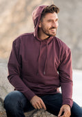 A bearded man sits outdoors on a rock, smiling to the side. He wears a maroon Woolx Chase Hoodie with the hood up and dark jeans. The softly blurred background adds to the relaxed vibe.