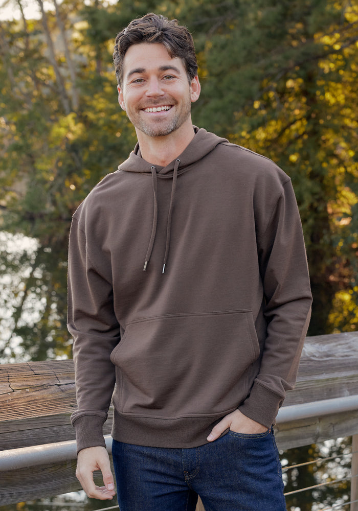 A man with short brown hair stands outdoors by a wooden railing, smiling in the Woolx Chase Hoodie • Final Sale - Bourbon Oak, paired with blue jeans, with trees and water visible in the background.