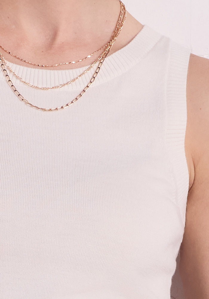 A close-up shows a person in the Chessy Tank Top - Dewdrop Pearl from Woolx, paired with two layered gold necklaces featuring flat chain links. The white background highlights only part of their neck and shoulder.
