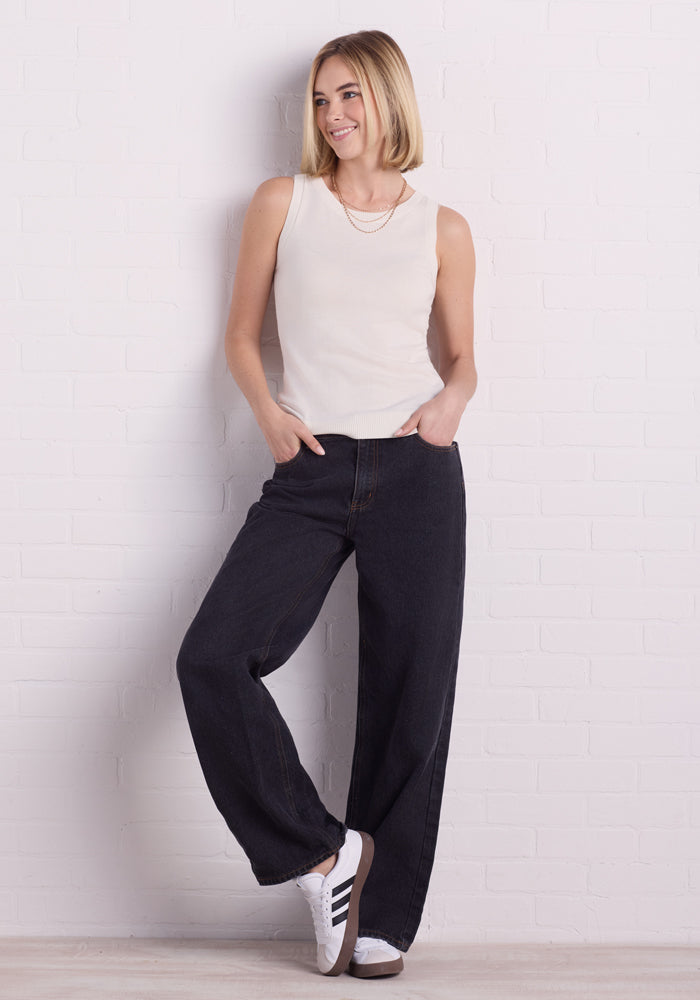 A woman with shoulder-length blonde hair leans against a white brick wall, wearing a Woolx Chessy Tank Top in Dewdrop Pearl, dark wide-leg jeans, white sneakers, and a necklace. Her hands are in her pockets as she slightly smiles while looking to the side.