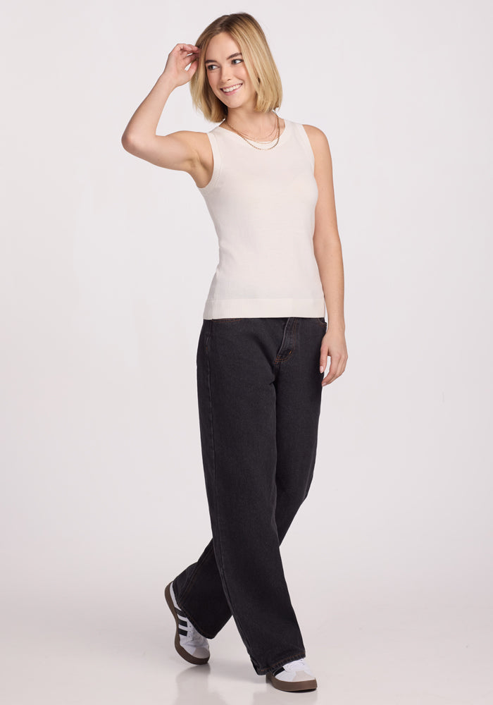 A person with short, blonde hair smiles while posing against a plain white background. They're wearing the Chessy Tank Top - Dewdrop Pearl by Woolx, black wide-leg pants, and white sneakers with black stripes. Their hand is near their head in the pose.