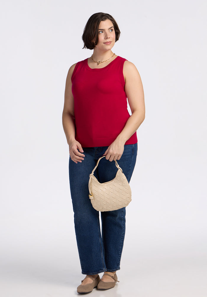 A woman with short brown hair wears the Woolx Chessy Tank Top in Poppy Spark, blue jeans, and beige shoes. She holds a small beige woven handbag and stands against a plain white background, looking slightly to her left.