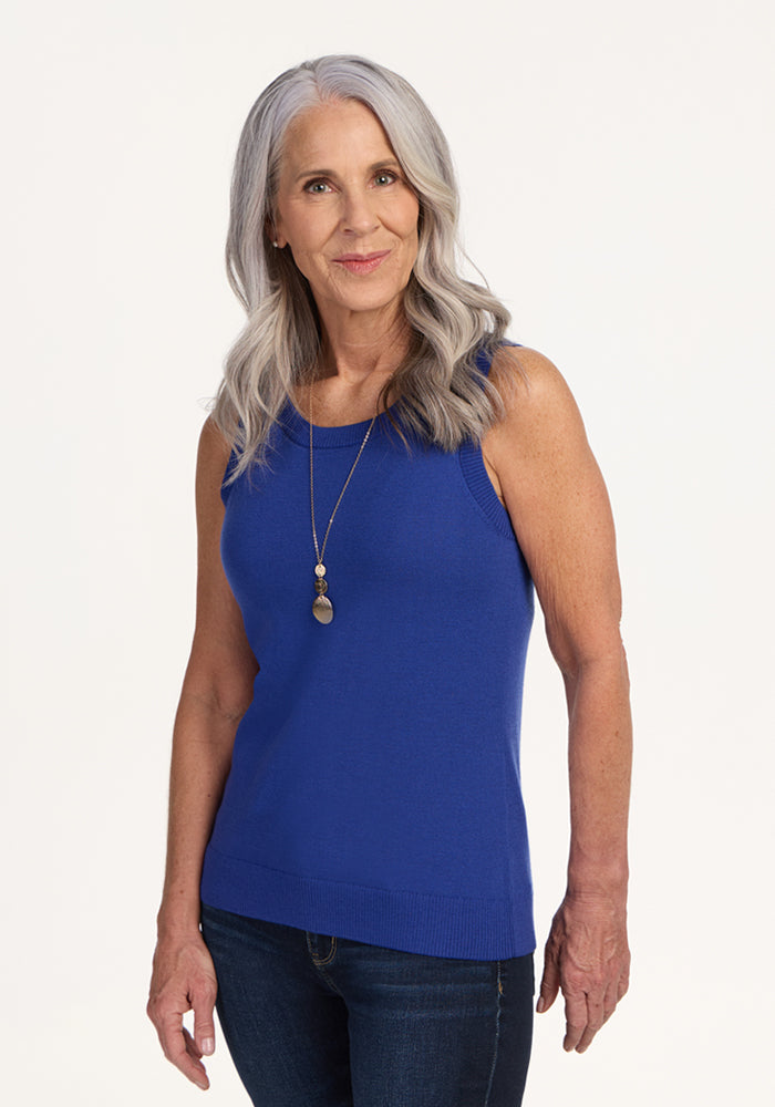 A smiling older woman with long gray hair wears the Woolx Chessy Tank Top in royal blue, paired with blue jeans and a long two-pendant necklace, standing against a plain light background. | Tam is 5’8.5”, wearing a size S