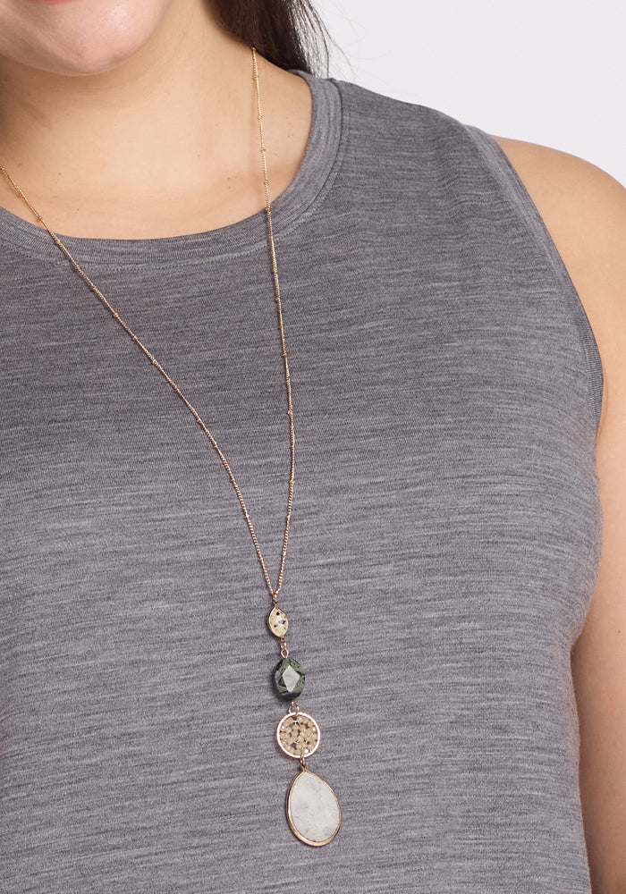 A woman is in a gray sleeveless top with a long gold necklace featuring diverse stone pendants, including white and speckled ones, exuding elegance. This ensemble pairs seamlessly with the Woolx Clara Dress in Graphite Heather, made from odor-resistant Merino wool for ultimate comfort.