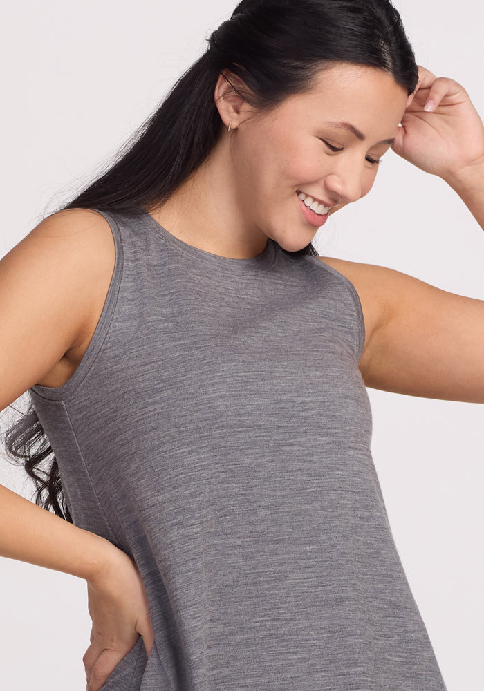 A person with long black hair smiles and looks down, posing in the sleeveless Clara Dress - Graphite Heather by Woolx. Their right hand touches their head, enjoying the comfort of its odor-resistant Merino wool material against a plain, light-colored background.