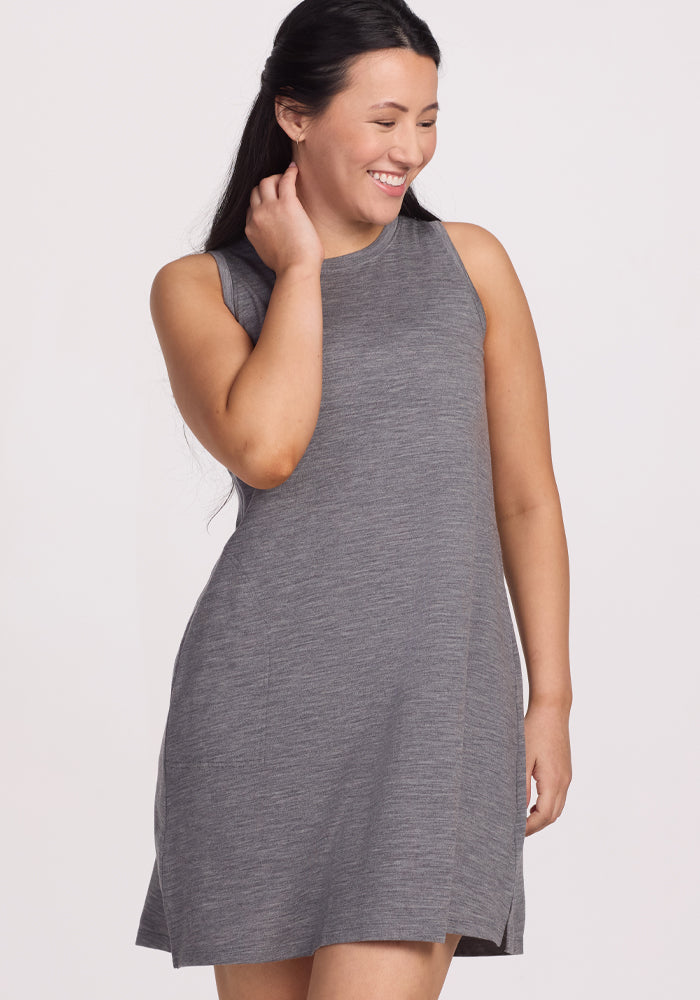 A woman with long dark hair smiles while wearing the sleeveless Clara Dress in Graphite Heather by Woolx, which features temperature-regulating Merino wool. She stands against a plain white background, gently touching her hair with one hand.