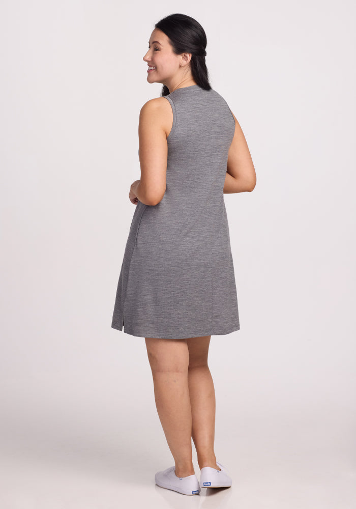 A person with long dark hair, smiling and hands clasped, stands against a plain white background. They wear the Woolx Clara Dress in Graphite Heather, a sleeveless Merino wool dress that's temperature-regulating and odor-resistant, paired with white shoes.