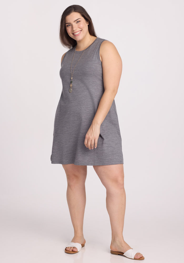 A smiling woman with long hair wears the sleeveless, temperature-regulating Clara Dress in Graphite Heather by Woolx and white sandals. She stands against a plain white background, accessorized with a long necklace. | Juliana is 5'10", wearing a size XL
