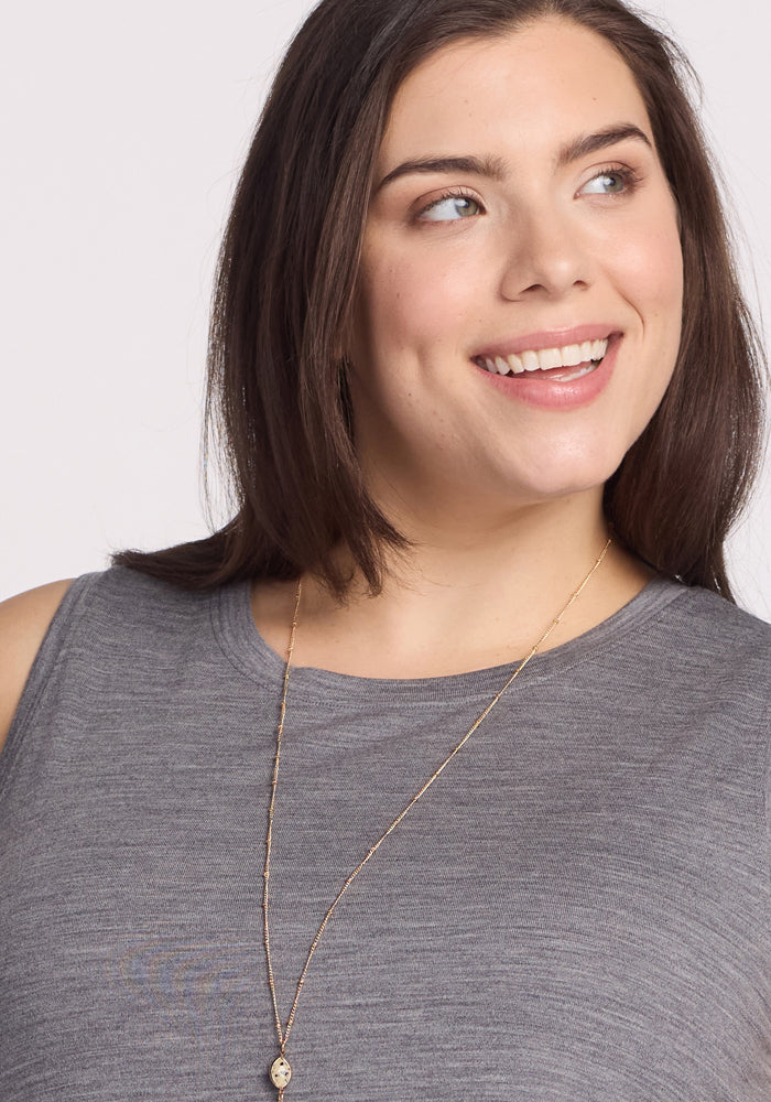 A smiling person with long brown hair wears the Clara Dress - Graphite Heather by Woolx. The sleeveless, breathable, and moisture-wicking dress pairs with a long necklace, enhancing their joyful expression as they look slightly to the side against a plain white background.