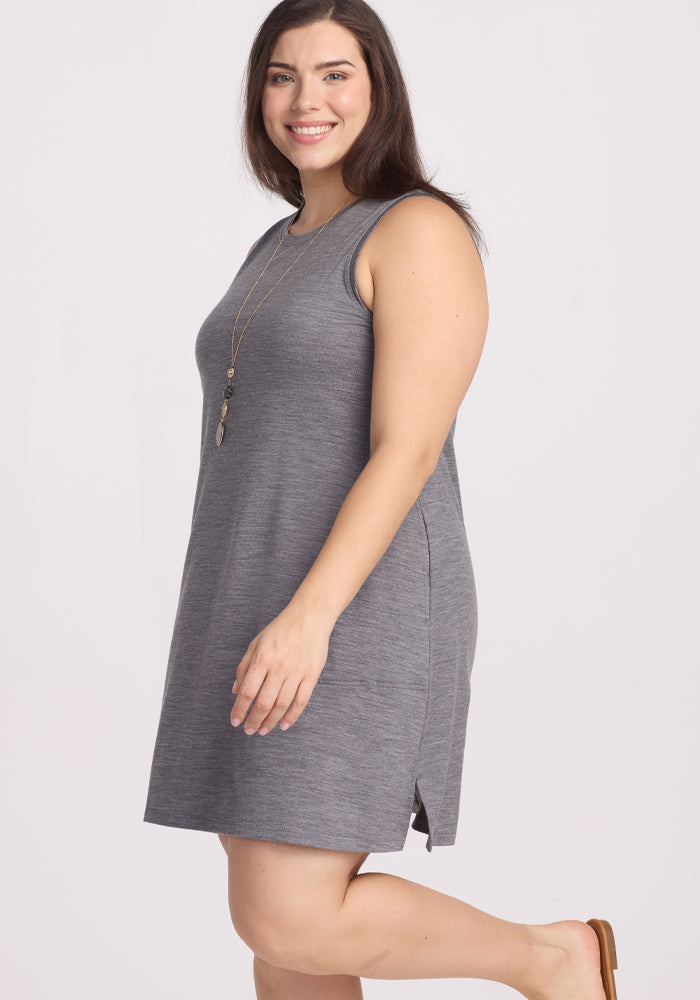 A person with long dark hair smiles while wearing a sleeveless Woolx Clara Dress in Graphite Heather, known for its odor resistance. They accessorize with a long necklace and stand sideways, gazing at the camera, set against a plain light-colored background.