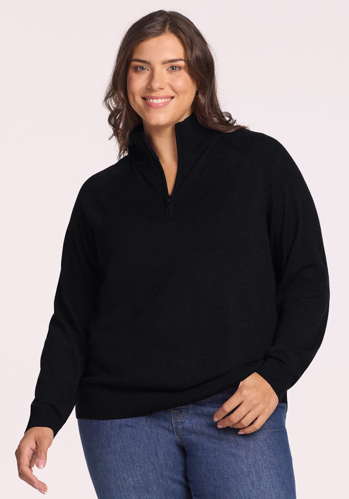A woman with medium-length brown hair smiles while wearing the Woolx Collins Half Zip Sweater in black and blue jeans, standing against a plain light background. | Juliana is 5'10", wearing a size XL