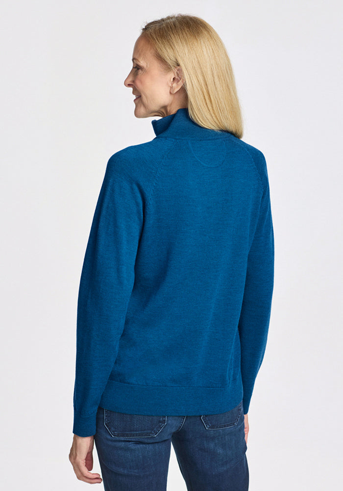 A woman with long blonde hair, wearing the Woolx Collins Half Zip Sweater in Moroccan Blue and jeans, stands facing away from the camera against a plain white background.