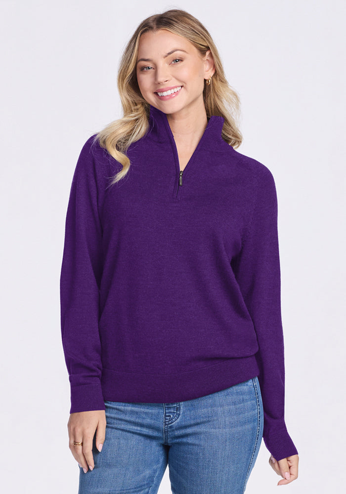 A woman with long, wavy blonde hair smiles while wearing the Woolx Collins Half Zip Sweater in Plum Crush and blue jeans, standing against a plain light background. | Saige is 5’7”, wearing a size XS