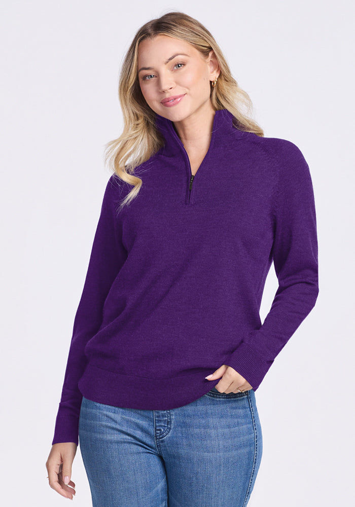 A woman with long blonde hair wears a Woolx Collins Half Zip Sweater in Plum Crush and blue jeans, standing against a plain light background and smiling gently at the camera.