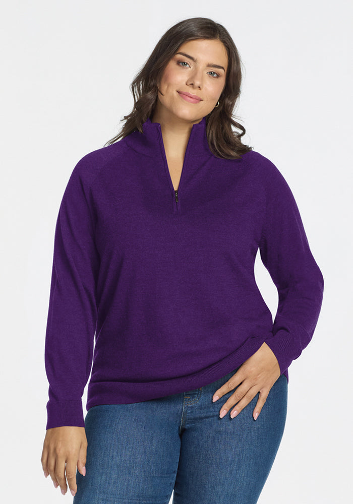 A woman with medium-length brown hair wears the Woolx Collins Half Zip Sweater in Plum Crush and blue jeans, standing against a plain white background and smiling softly with one hand on her hip. | Juliana is 5'10", wearing a size XL