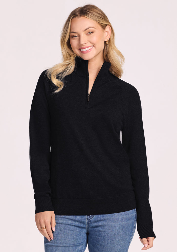 A woman with long blonde hair smiles, wearing the Woolx Collins Half Zip Sweater in black and blue jeans, standing against a plain light background. | Saige is 5'7", wearing a size XS