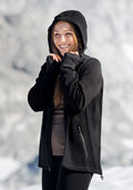 A smiling woman wears the Woolx Cubby Hooded Sweatshirt in black, standing outdoors in a snowy mountain landscape with her hands near her face, enjoying the cold weather.