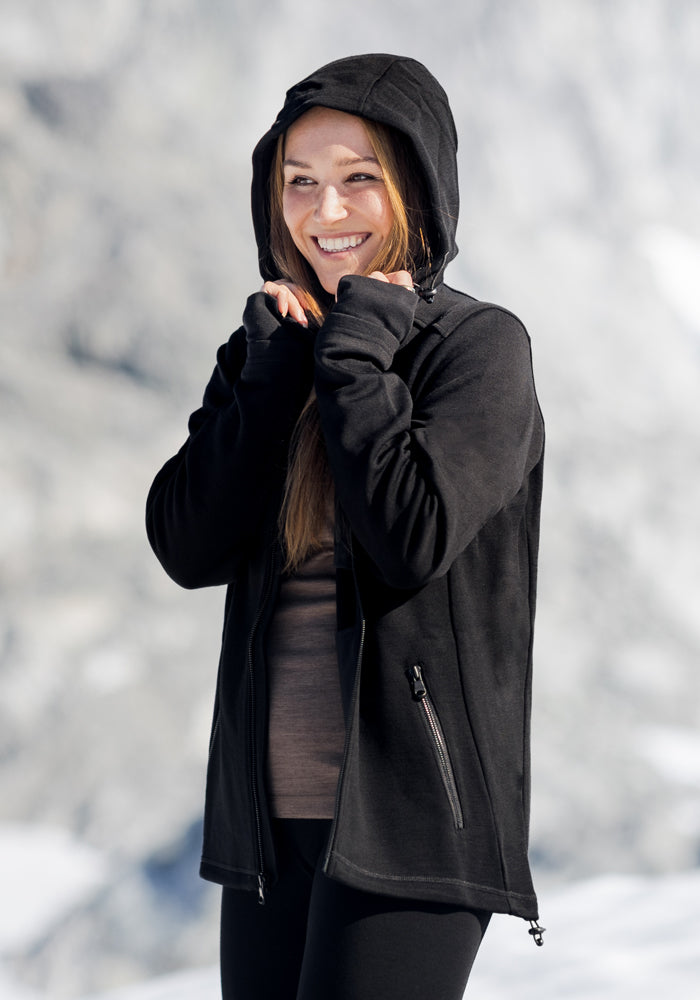 A smiling woman wears the Woolx Cubby Hooded Sweatshirt in black, standing outdoors in a snowy mountain landscape with her hands near her face, enjoying the cold weather.
