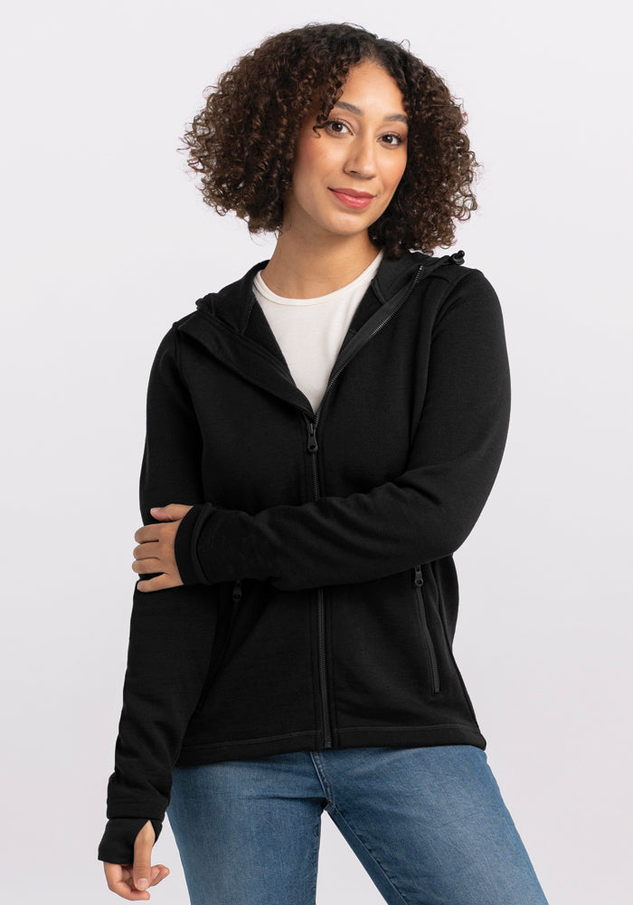 A person with curly hair is wearing a Woolx Cubby Hooded Sweatshirt in black with heavy-duty zippers over a white shirt and blue jeans. They are standing against a plain light-colored background, with one arm crossing their body and the other hanging by their side. | Tori is 5'7", wearing a size S