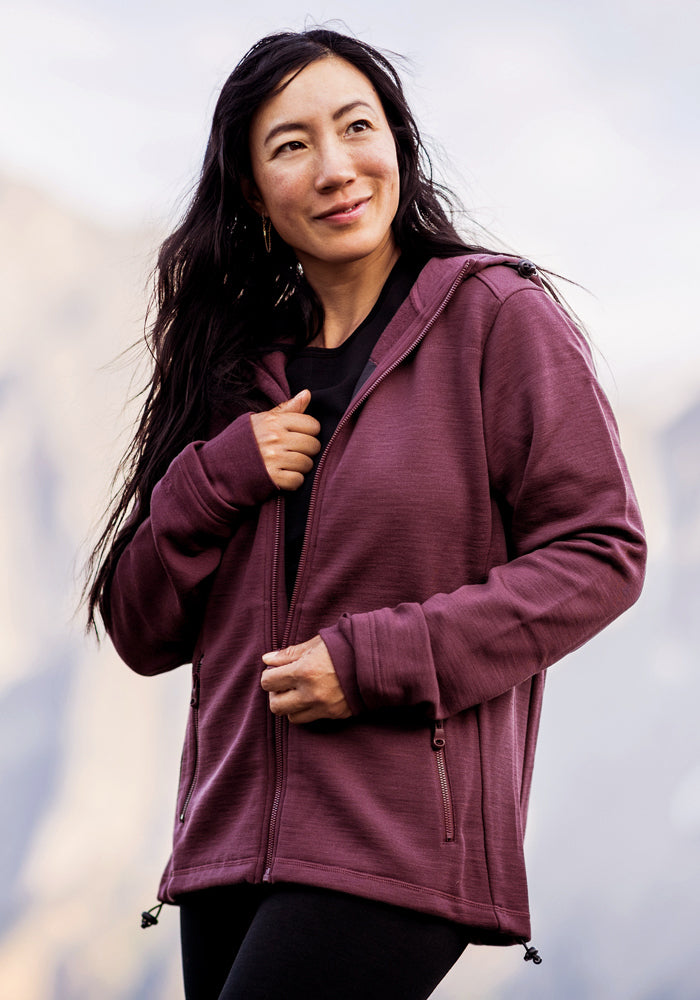 A woman with long dark hair smiles while wearing the Woolx Cubby Hooded Sweatshirt in Grape Harvest, standing outdoors against a blurred, natural background.