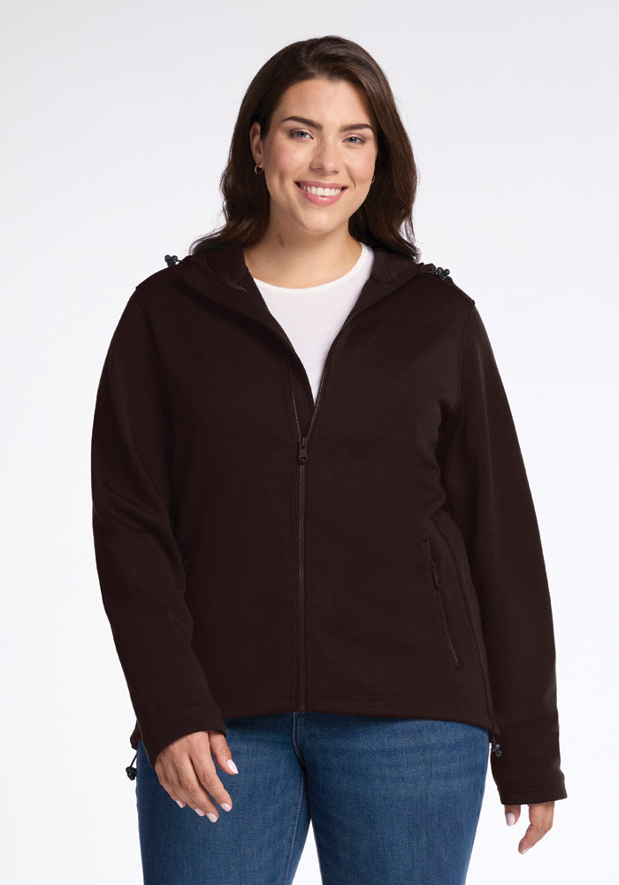 A person with long hair smiles, wearing a Woolx Cubby Hooded Sweatshirt in French Roast over a white shirt and blue jeans against a plain white background.