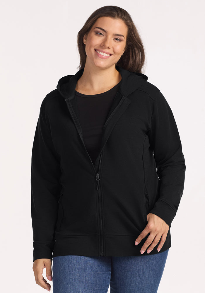 A woman with long brown hair smiles wearing the Woolx Darby Zip Up Hoodie in black, paired with a black shirt and blue jeans, standing against a plain light background. | Juliana is 5'10", wearing a size XL