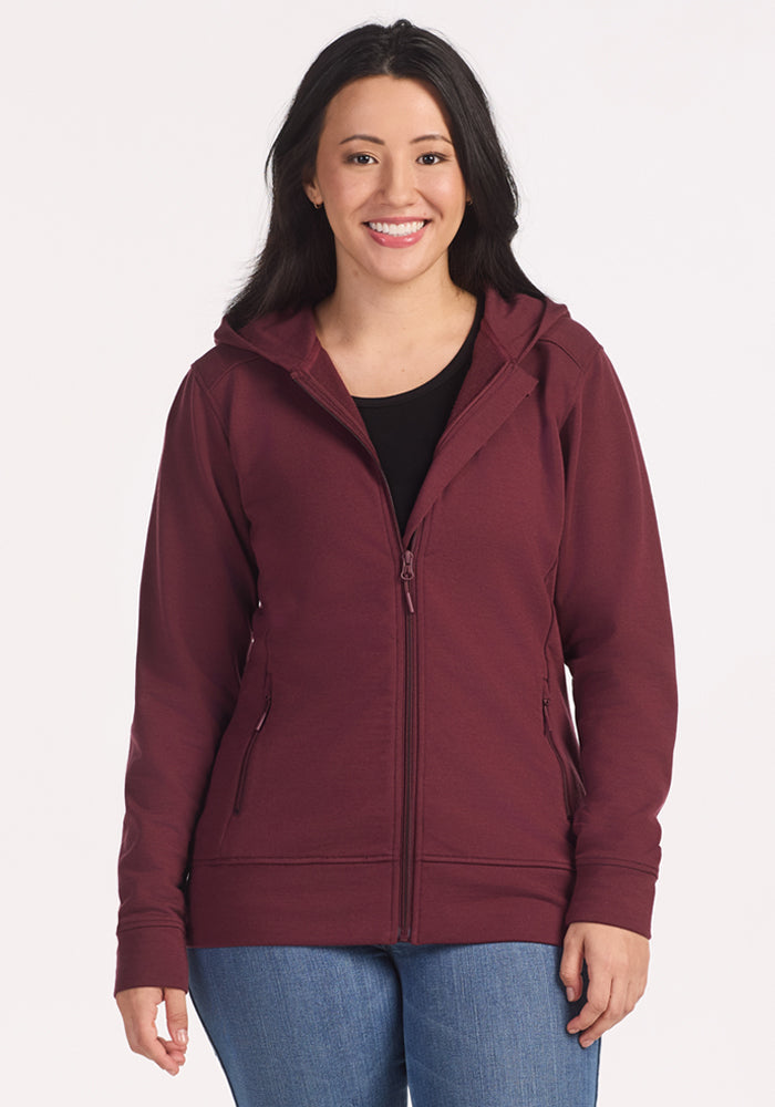 A woman with long dark hair smiles while wearing the Woolx Darby Zip Up Hoodie in Toasted Garnet over a black top and blue jeans, standing against a plain white background.