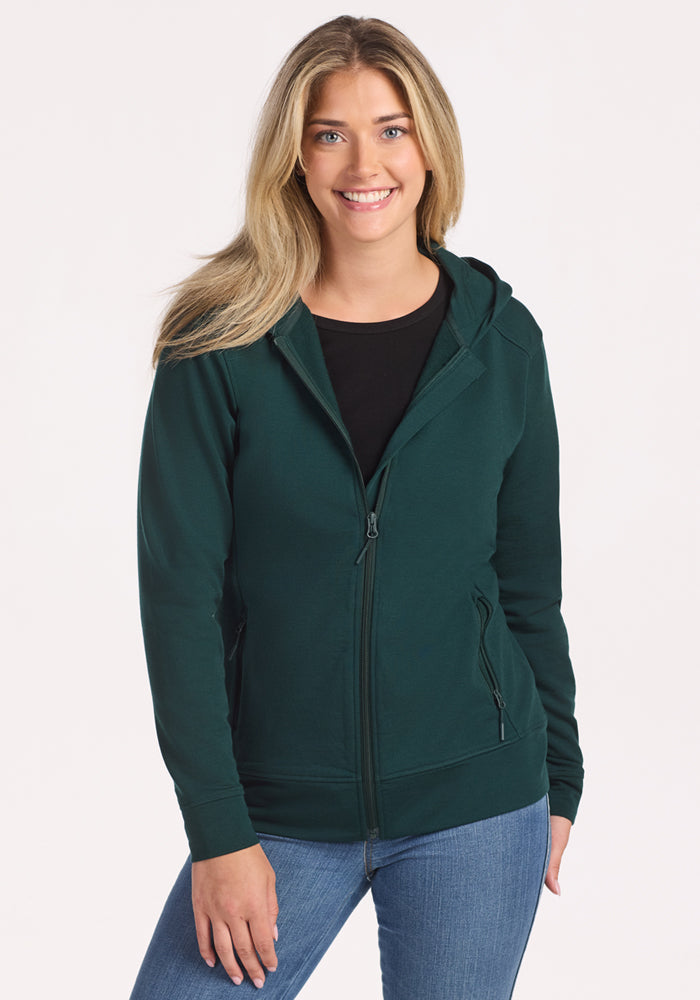 A smiling woman with long blonde hair wears the Woolx Darby Zip Up Hoodie in Deep Timber over a black shirt and blue jeans, standing against a plain white background.