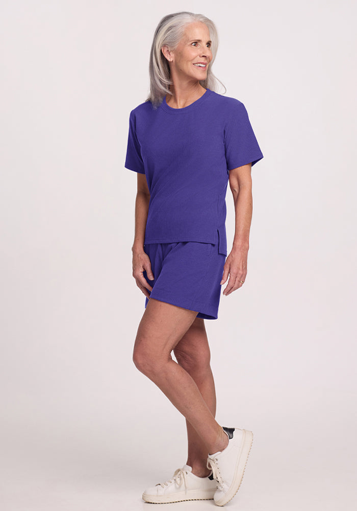 Older woman wearing Woolx Delia Ribbed Short Sleeve in Berry Indigo, shorts, and white sneakers, standing and smiling.