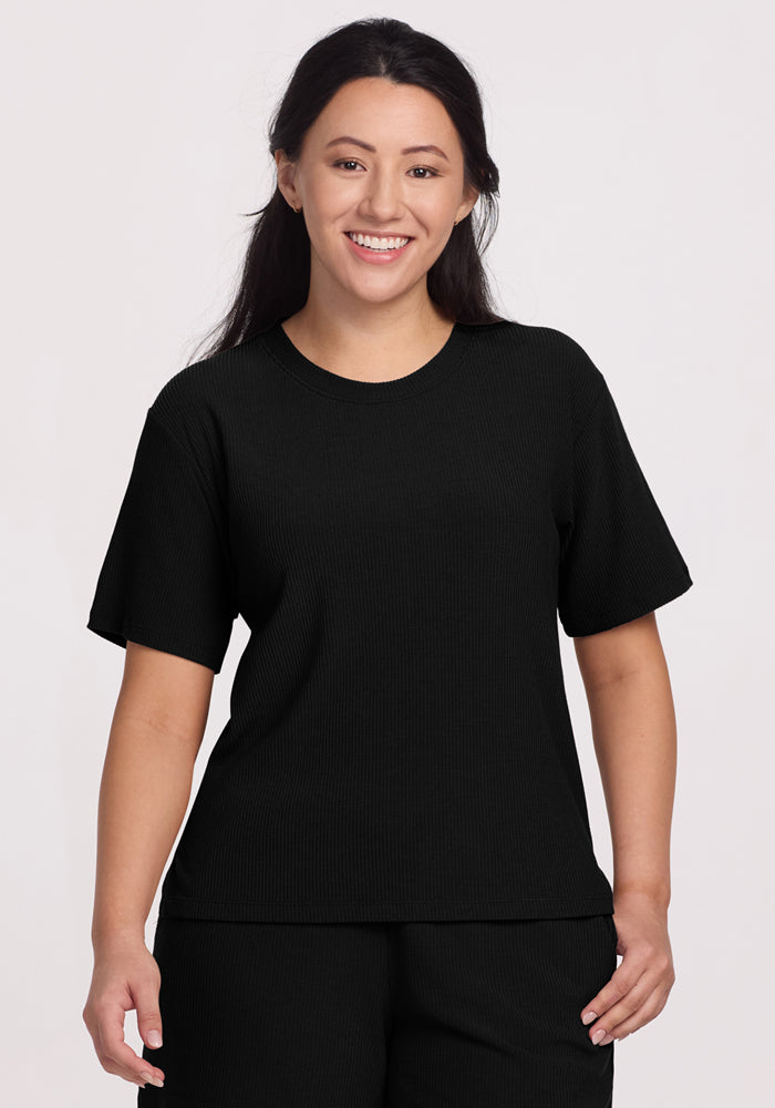 A person with long dark hair smiles, wearing the luxurious Woolx Delia Ribbed Top in black, paired with matching pants. The temperature-regulating outfit stands out against the plain, light-colored background. | Sarah is 5’8”, wearing a size S