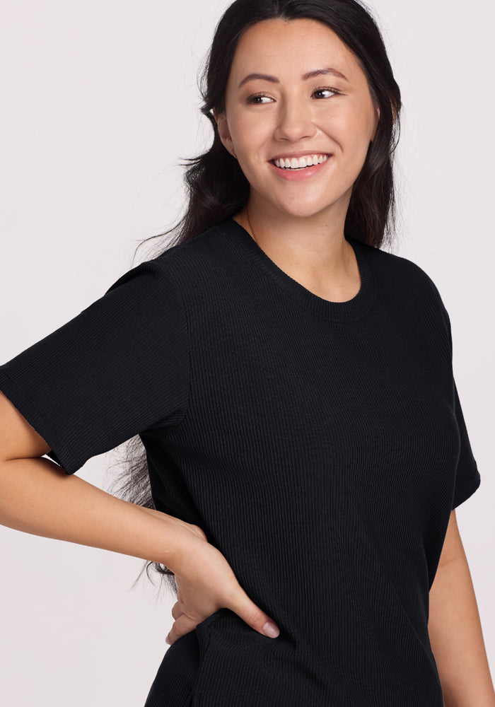 A person with long dark hair, wearing the Woolx Delia Ribbed Short Sleeve in black, smiles while looking to the side. They have one hand on their hip against a plain white background.