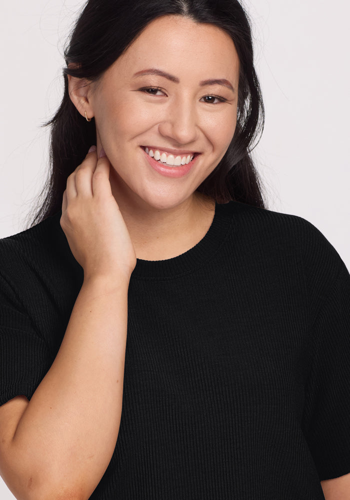 A person with long dark hair smiles warmly, touching their ear. They wear the Woolx Delia Ribbed Short Sleeve in black, set against a light background to highlight the elegance of its design.