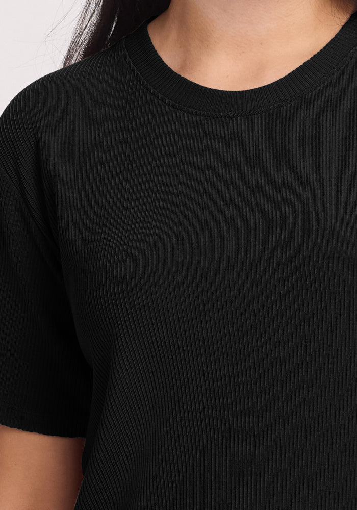 A person wears the Woolx Delia Ribbed Short Sleeve in black, highlighting the rich texture of its fabric, short sleeves, and round neckline. The plain background accentuates the elegant simplicity of this classic piece.