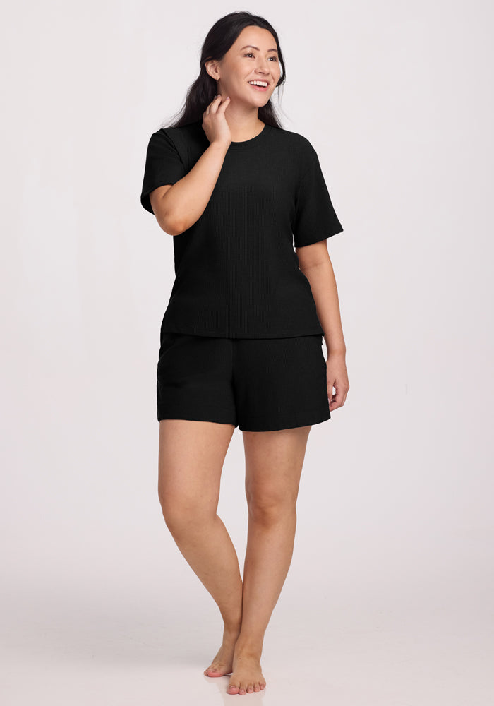 A person with long dark hair, barefoot and smiling, touches their face while wearing the Delia Ribbed Short Sleeve in black by Woolx and matching shorts against a plain background.
