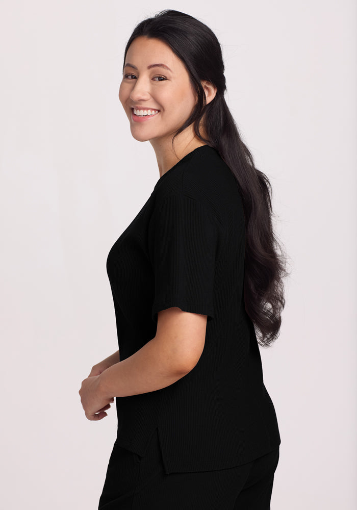 A smiling woman with long dark hair wears the Woolx Delia Ribbed Short Sleeve in black, its loose fit enhancing her relaxed vibe. She gazes to the side, set against a plain white background.