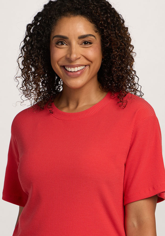 A woman with curly hair smiles in the Woolx Delia Ribbed Short Sleeve - Hibiscus against a plain background.