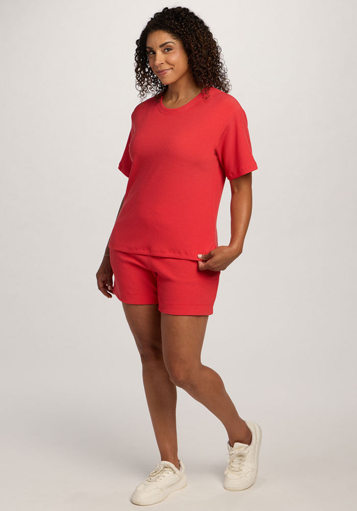 Woman smiling in the Woolx Delia Ribbed Short Sleeve - Hibiscus and red shorts, paired with white sneakers.