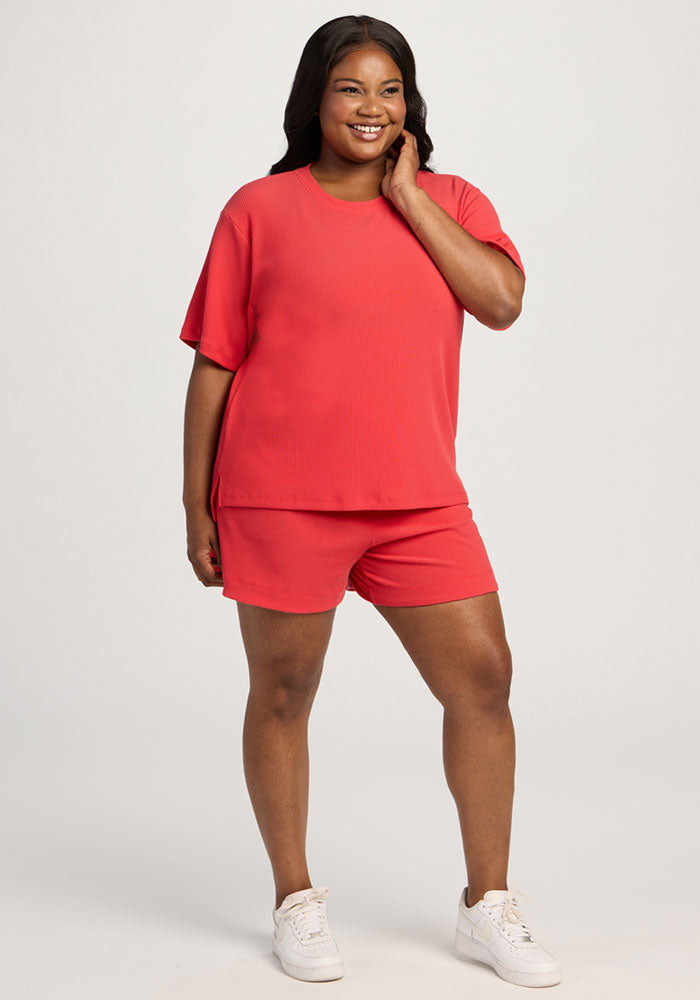 A woman wears the Woolx Delia Ribbed Short Sleeve - Hibiscus with matching shorts and white sneakers, smiling.