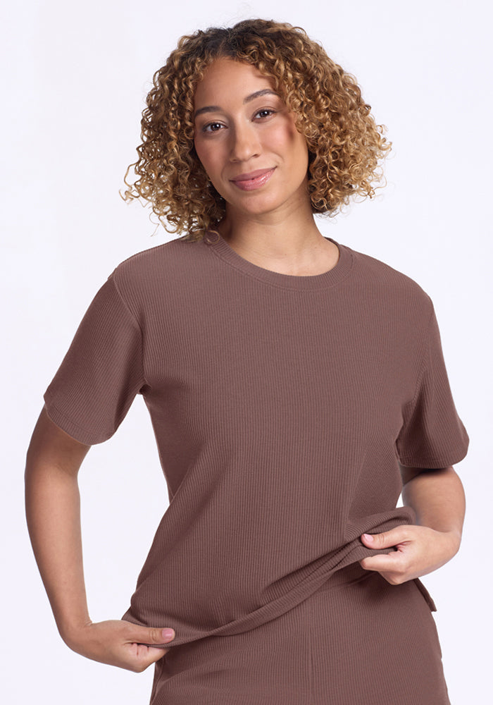 Smiling person with curly hair in Woolx Delia Ribbed Short Sleeve in Peppercorn, holding the shirt's hem. | Tori is 5'7", wearing a size S