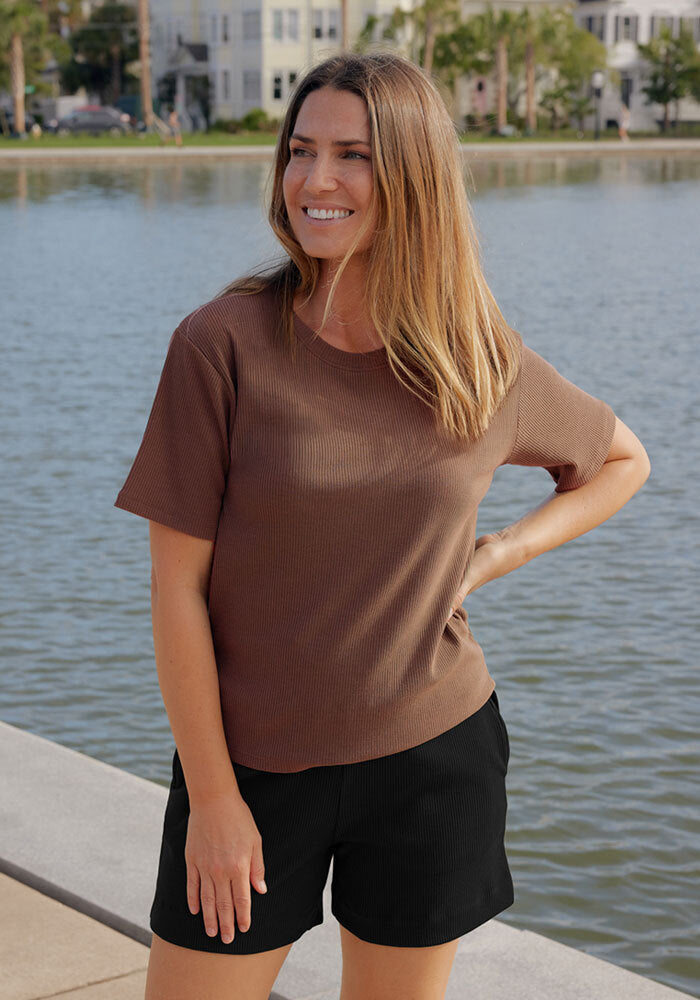 A woman in a Woolx Delia Ribbed Short Sleeve - Sienna Kiss smiles by the waterfront, one hand on her hip.