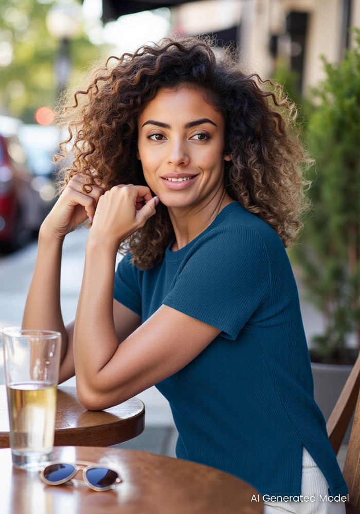 A woman in a Woolx Delia Ribbed Short Sleeve – Moroccan Blue, sits smiling at an outdoor café with a drink and sunglasses.