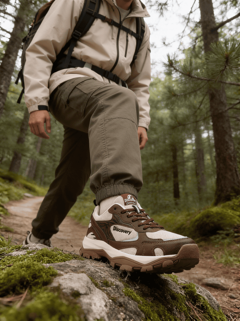 Person hiking in a forest wearing a jacket, pants, and a backpack with a focus on a brown and white hiking shoe.