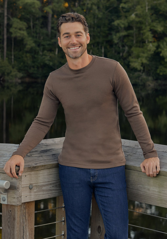 A man with short brown hair and a beard, wearing the Woolx Easton Crew Neck Shirt in Bourbon Oak and blue jeans, smiles while standing on a wooden deck beside a lake with trees in the background.