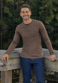 A man with short brown hair and a beard, wearing the Woolx Easton Crew Neck Shirt in Bourbon Oak and blue jeans, smiles while standing on a wooden deck beside a lake with trees in the background.